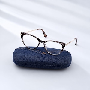 May include: Cat-eye glasses with a brown and black tortoiseshell pattern on the frames and thin gold-tone metal temples. The glasses are resting on top of a dark blue denim-textured hard-shell case with visible stitching details along the top.