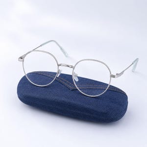May include: Round silver-toned metal eyeglasses with clear lenses resting on a dark blue denim-textured hard shell case. The glasses feature thin wire frames, a straight bridge, and slim temples with light-colored tips, presented as a set.