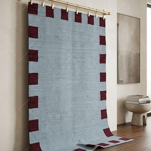 Peut inclure: Un tapis tissé rectangulaire bleu clair avec une bordure marron foncé présentant un motif de carrés répétitifs. Le tapis est suspendu à une tringle en métal doré par une série de pinces métalliques assorties, tombant verticalement.
