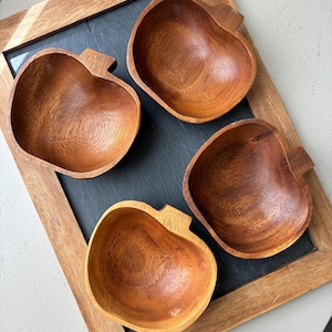 May include: Four apple-shaped wooden bowls with a smooth, polished finish and visible natural wood grain patterns. Each bowl features a small, rectangular stem-like protrusion at the top edge, arranged on a dark gray rectangular slate board.