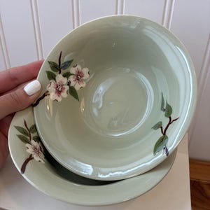 Może przedstawiać: Zestaw dwóch jasnych, szałwiowo-zielonych ceramicznych misek z delikatnym, ręcznie malowanym wzorem brązowych gałązek z zielonymi liśćmi oraz białymi kwiatami z różowymi środkami. Miski mają gładkie, błyszczące wykończenie i zaokrąglony kształt.
