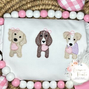 May include: Three embroidered dogs holding hearts, each dog is a different color and breed. The dogs are on a white shirt with the text "Brownie Points Boutique Embroidery & Applique" in the bottom right corner.