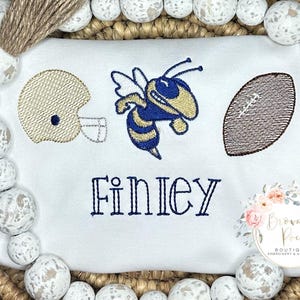 May include: White onesie with embroidered football, bee, and helmet in gold and blue. The name "Finley" is embroidered in blue below the sports imagery. A small floral logo is in the bottom right corner.
