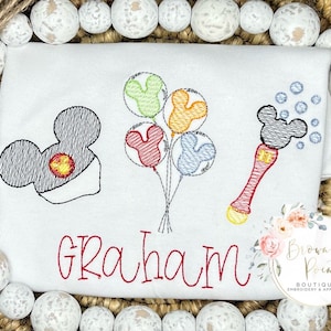May include: A white embroidered t-shirt with a Mickey Mouse head, a bunch of colorful balloons, a magic wand with Mickey Mouse ears, and the name "Graham" embroidered in red.