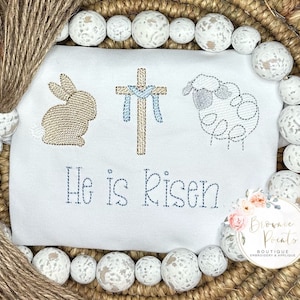 May include: White embroidered baby onesie with a brown bunny, a cross with blue ribbon, and a gray lamb. The onesie has the text "He is Risen" embroidered in brown thread.