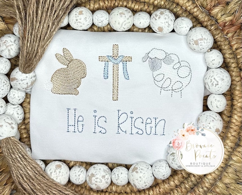 May include: White embroidered baby onesie with a brown bunny, a cross with blue ribbon, and a gray lamb. The onesie has the text "He is Risen" embroidered in brown thread.