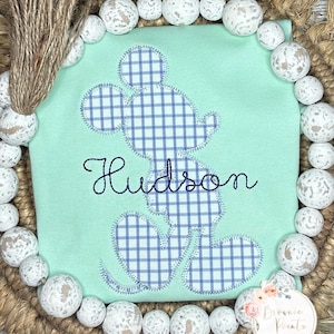 May include: A light green t-shirt with a blue and white plaid Mickey Mouse applique. The name "Hudson" is embroidered in black below the applique.
