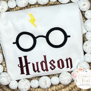 May include: White t-shirt with a black embroidered pair of glasses, a yellow lightning bolt, and the name "Hudson" in maroon script.