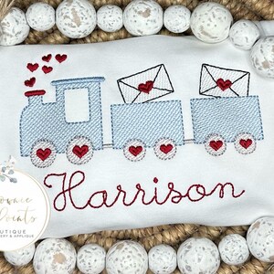 May include: A white embroidered shirt with a blue train carrying red hearts and envelopes. The name "Harrison" is embroidered in red below the train.