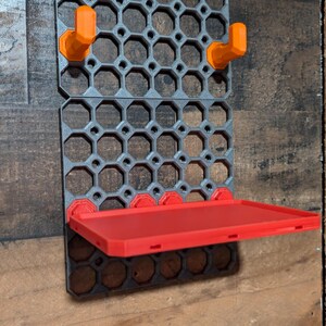 May include: A gray 3D-printed wall organizer with a honeycomb pattern, featuring orange hooks and a red shelf. The organizer is mounted on a wooden surface, providing a storage solution.