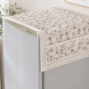 May include: A beige refrigerator cover featuring a dark brown floral pattern and intricate white lace trim along the edges. The cover includes side pockets for storage and drapes over the top and sides of a light grey refrigerator unit.
