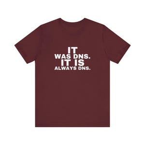 Peut inclure: Un t-shirt à manches courtes bordeaux avec un texte blanc centré sur la poitrine qui indique : IT WAS DNS. IT IS ALWAYS DNS. Le vêtement possède une encolure ronde et une coupe standard, fabriqué dans un tissu uni à la texture lisse.