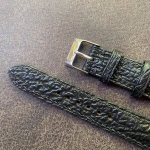 K&ouml;nnte beinhalten: Nahaufnahme eines schwarzen Uhrenarmbands mit strukturierter Oberfl&auml;che und silberner Schnalle. Das Armband hat eine Reihe von L&ouml;chern zur Anpassung und ist mit einem Kontrastfaden gen&auml;ht. Das Uhrenarmband ist aus Leder.
