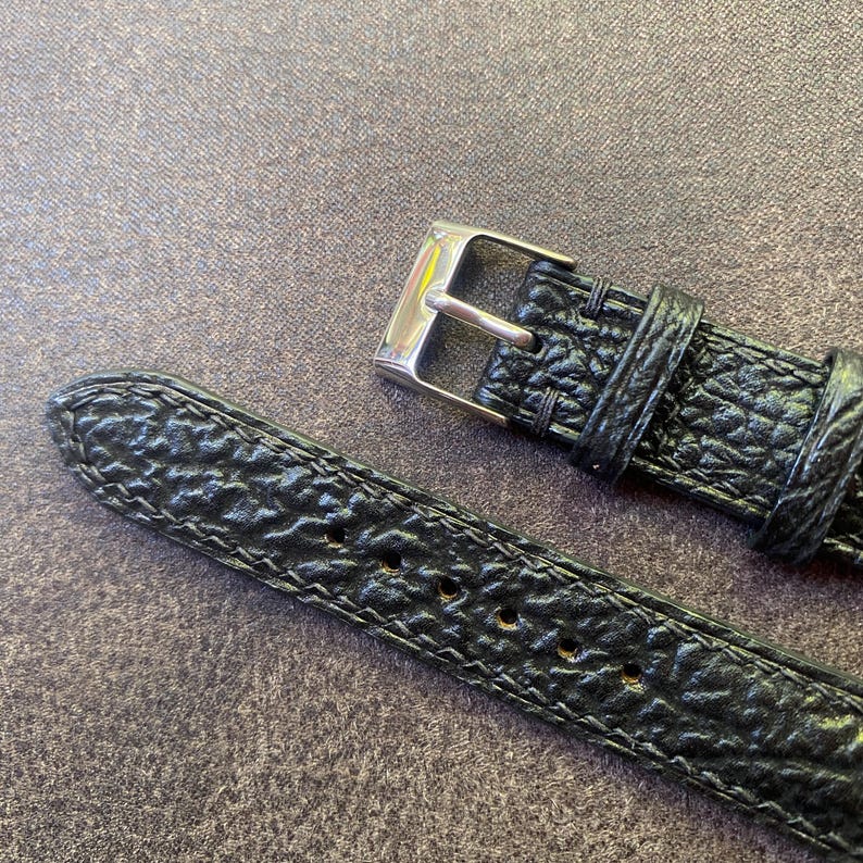 K&ouml;nnte beinhalten: Nahaufnahme eines schwarzen Uhrenarmbands mit strukturierter Oberfl&auml;che und silberner Schnalle. Das Armband hat eine Reihe von L&ouml;chern zur Anpassung und ist mit einem Kontrastfaden gen&auml;ht. Das Uhrenarmband ist aus Leder.