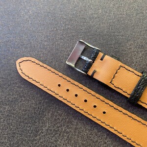 K&ouml;nnte beinhalten: Nahaufnahme eines Uhrenarmbands. Das Armband ist hellbraun mit schwarzen N&auml;hten und einem schwarzen Lederabschnitt in der N&auml;he der Edelstahlschnalle. Das Armband hat mehrere L&ouml;cher zur Anpassung.