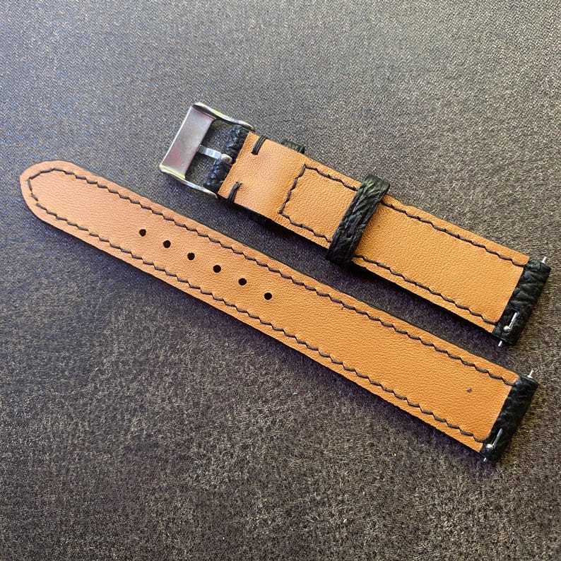 K&ouml;nnte beinhalten: Ein Uhrenarmband mit hellbraunem Innen- und schwarzem Au&szlig;enbereich. Das Armband hat schwarze N&auml;hte und eine silberne Schnalle. Das Armband ist aus Leder und hat ein klassisches Design.