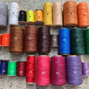 K&ouml;nnte beinhalten: Eine Sammlung von Garnrollen in verschiedenen Farben, darunter Grau, Wei&szlig;, Schwarz, Gelb, Orange, Braun, Blau, Gr&uuml;n, Rot und Lila. Die Rollen sind von 1 bis 27 nummeriert.