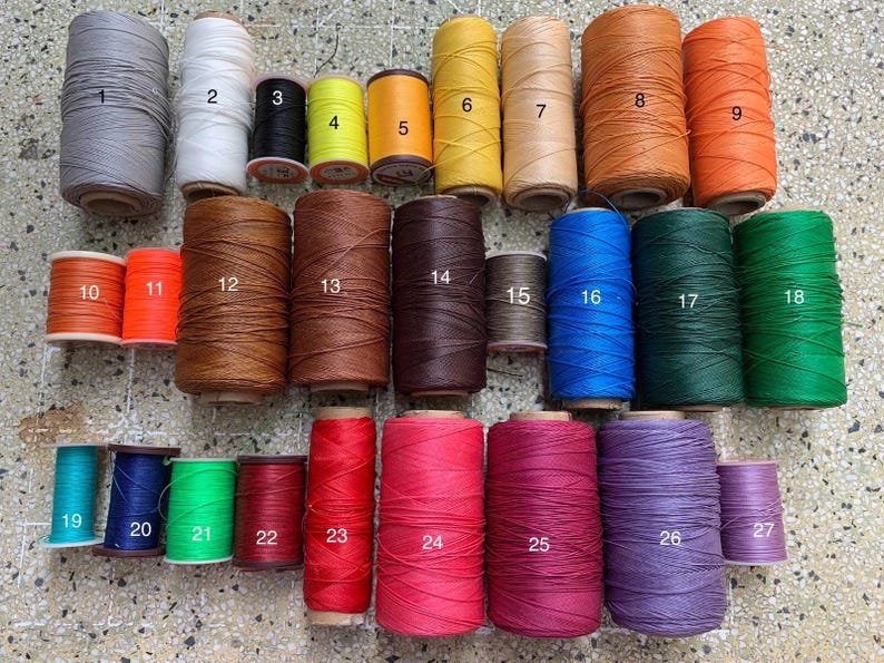 K&ouml;nnte beinhalten: Eine Sammlung von Garnrollen in verschiedenen Farben, darunter Grau, Wei&szlig;, Schwarz, Gelb, Orange, Braun, Blau, Gr&uuml;n, Rot und Lila. Die Rollen sind von 1 bis 27 nummeriert.