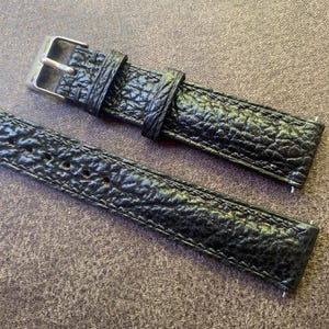 K&ouml;nnte beinhalten: Nahaufnahme eines schwarzen Uhrenarmbands mit einem strukturierten, gepr&auml;gten Muster. Das Armband verf&uuml;gt &uuml;ber eine silberfarbene Schnalle und eine passende Schlaufe. Das Lederarmband ist an den R&auml;ndern gen&auml;ht.