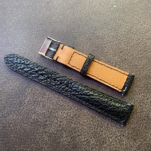 K&ouml;nnte beinhalten: Schwarzes Uhrenarmband mit strukturierter Oberfl&auml;che und Edelstahlschnalle. Das Armband hat eine hellbraune Unterseite und schwarze N&auml;hte. Das Uhrenarmband wird auf einer dunkelgrauen Oberfl&auml;che gezeigt.