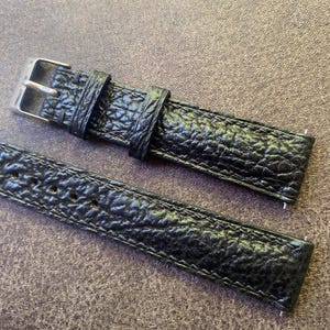 K&ouml;nnte beinhalten: Nahaufnahme eines schwarzen Uhrenarmbands mit strukturierter, gemusterter Oberfl&auml;che. Das Armband verf&uuml;gt &uuml;ber eine silberfarbene Schnalle und eine passende Schlaufe. Das Uhrenarmband besteht aus einem strapazierf&auml;higen Material.