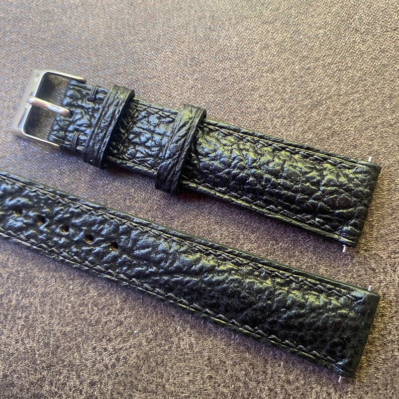 K&ouml;nnte beinhalten: Nahaufnahme eines schwarzen Uhrenarmbands mit strukturierter, gemusterter Oberfl&auml;che. Das Armband verf&uuml;gt &uuml;ber eine silberfarbene Schnalle und eine passende Schlaufe. Das Uhrenarmband besteht aus einem strapazierf&auml;higen Material.