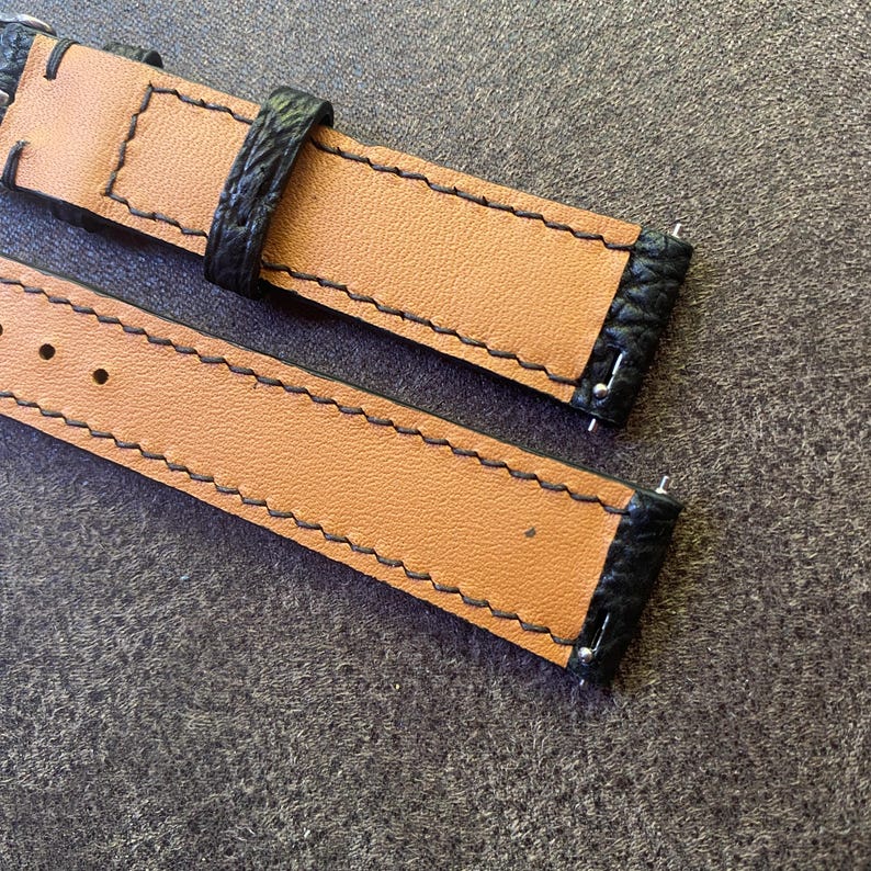 K&ouml;nnte beinhalten: Nahaufnahme von zwei Uhrenarmb&auml;ndern. Die Armb&auml;nder bestehen aus hellbraunem Leder mit schwarzen Kanten und N&auml;hten. Die schwarzen Kanten haben ein strukturiertes Aussehen. Die Armb&auml;nder liegen auf einer grauen Oberfl&auml;che.