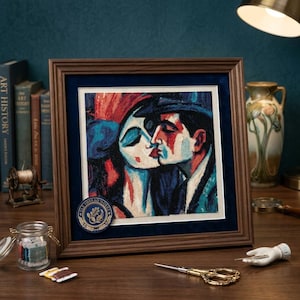 May include: A framed needlepoint artwork depicting a couple kissing in a stylized manner with bold blue, red, and black colors. The square piece is set in a dark wood frame with a navy blue mat and a circular emblem labeled Rae Thread Works.