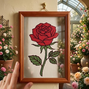 May include: A framed cross-stitch of a red rose with green leaves and stem. The artwork is in a wooden frame and hangs on a beige wall. The background shows a greenhouse with potted roses.