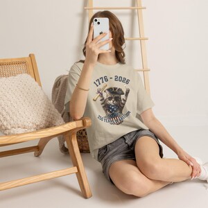 May include: A beige t-shirt featuring a graphic of a cat wearing a tricorn hat and sunglasses, holding a scroll. The text reads 1776-2026 and 250 YEARS OF FREEDOM, with a patriotic shield design below the cat, showcasing a red, white, and blue theme.
