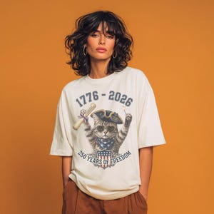 May include: An off-white t-shirt featuring a graphic of a cat wearing a tricorn hat and sunglasses, holding a scroll and a sparkler. The text reads 1776 - 2026 and 250 YEARS OF FREEDOM, with a shield displaying the American flag design below.