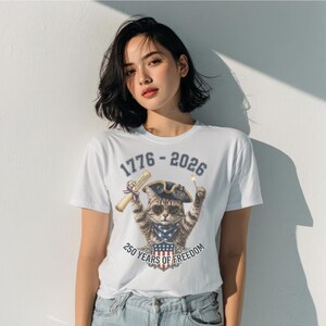 May include: A white t-shirt featuring a graphic of a tabby cat wearing a colonial hat and heart-shaped sunglasses. The cat holds a scroll and a sparkler. Text reads 1776-2026 and 250 YEARS OF FREEDOM above and below the cat illustration.