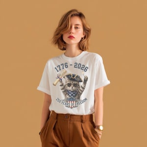 May include: A white t-shirt featuring a graphic of a cat wearing a tricorn hat, sunglasses, and a star-spangled bandana. The cat holds a scroll and a sparkler. Text reads 1776-2026 and 250 YEARS OF FREEDOM above and below the cat illustration.