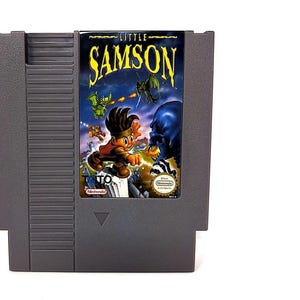 May include: A gray Nintendo Entertainment System (NES) game cartridge for "Little Samson." The label features a cartoon illustration of a boy fighting monsters, with the game title in yellow. The cartridge is rectangular with a textured design.