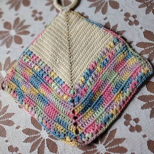 May include: A handmade, square crochet potholder with a loop for hanging. The center is cream-colored, with a chevron pattern. The border features stripes of pink, blue, yellow, and green. The potholder is designed for kitchen use.