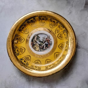 May include: A decorative, vintage plate with a gold rim and a yellow center. The center features a small, detailed painting of two figures in a garden setting. The plate is adorned with gold filigree patterns.