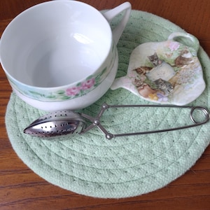 May include: A white teacup with a floral design and a stainless steel tea infuser rest on a light green coaster. A decorative saucer with a cat motif is also present. The teacup has a pink rose pattern.