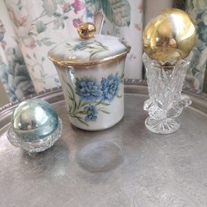 May include: A silver serving tray holds a small, ornate glass vase with a gold sphere, a small blue and silver orb, and a white and gold floral jar with a lid and spoon. The jar features blue flower illustrations.
