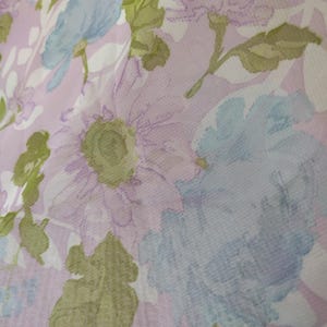 May include: Close-up of a fabric with a floral pattern. The design features large, soft-focus flowers in shades of lavender, light blue, and green against a pale pink background. The fabric has a delicate, slightly sheer appearance.