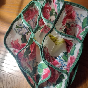 May include: A fabric organizer with multiple compartments featuring a white background with a floral pattern of red roses, green leaves, and small blue flowers. The edges of each section are finished with a dark green fabric trim.