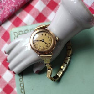 May include: A gold-tone wristwatch with a round, cream-colored analog face featuring black Arabic numerals and thin black hands. The watch has a metallic expansion band with engraved detailing on the links connecting to the watch case.