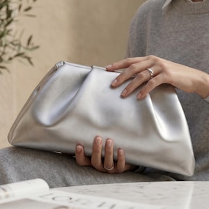 May include: A metallic silver clutch bag with a soft, gathered, and pleated design. The pouch features a smooth, reflective surface and a structured frame closure, presenting a sleek, minimalist aesthetic suitable for carrying essential items.