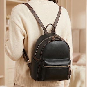 May include: A black backpack with a rounded top, a front zippered pocket, and brown patterned shoulder straps. The bag features gold-toned metal zippers and a top handle, with a textured finish on the main body and matching brown trim details.