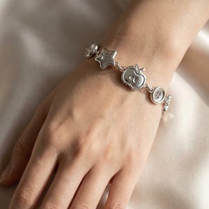 May include: Silver charm bracelet with star, apple, and button charms. The bracelet is worn on a hand, with the charms linked together. The bracelet is made of a shiny metal and has a delicate design. The bracelet is approximately 18 cm in length.