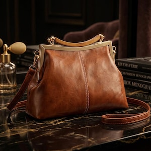 Könnte beinhalten: Eine braune Ledertasche mit einem strukturierten goldfarbenen Metallrahmen, einem geschwungenen Holzgriff an der Oberseite und einem abnehmbaren Schulterriemen. Die Tasche hat eine glatte, polierte Oberfläche mit sichtbaren Nähten und einem Verschluss.