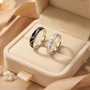 May include: Two matching silver rings with a swirling design, one black and one white, displayed in a beige ring box. The rings are adjustable. The background includes a candle, flowers, and satin fabric.