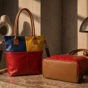 Peut inclure: Un sac cabas color-block composé de panneaux de cuir bleu marine, jaune et rouge avec des anses en cuir beige et une bandoulière amovible. Un second sac assorti est posé horizontalement, révélant sa base et ses côtés en cuir beige.