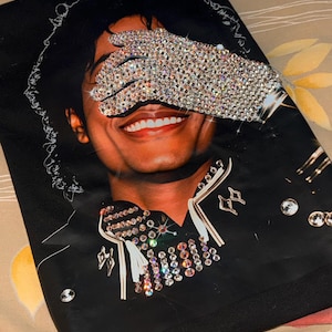 May include: Black graphic t-shirt featuring a smiling person with a sparkling glove covering their eyes. The shirt has a detailed design with rhinestones and embellishments. The image is a portrait style.
