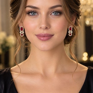 May include: A pair of drop earrings featuring a rectangular red gemstone surrounded by a halo of small clear stones, connected to a smaller circular cluster of clear stones set in a gold-toned metal frame, worn on the earlobes.