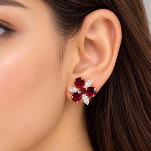 Puede incluir: Un pendiente de botón en tono dorado con un diseño floral compuesto por tres piedras de color rojo intenso en talla pera y dos piedras blancas transparentes en talla marquesa, engastadas en un marco metálico con forma de flor.